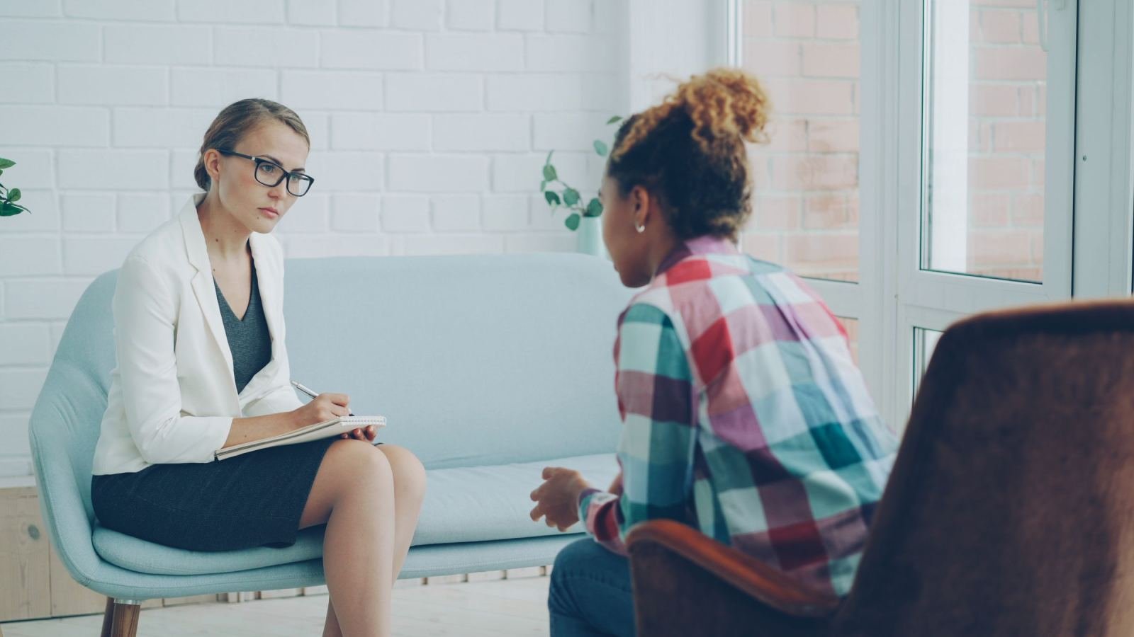 A counselor in a white blazer takes notes while talking to a client in a plaid shirt