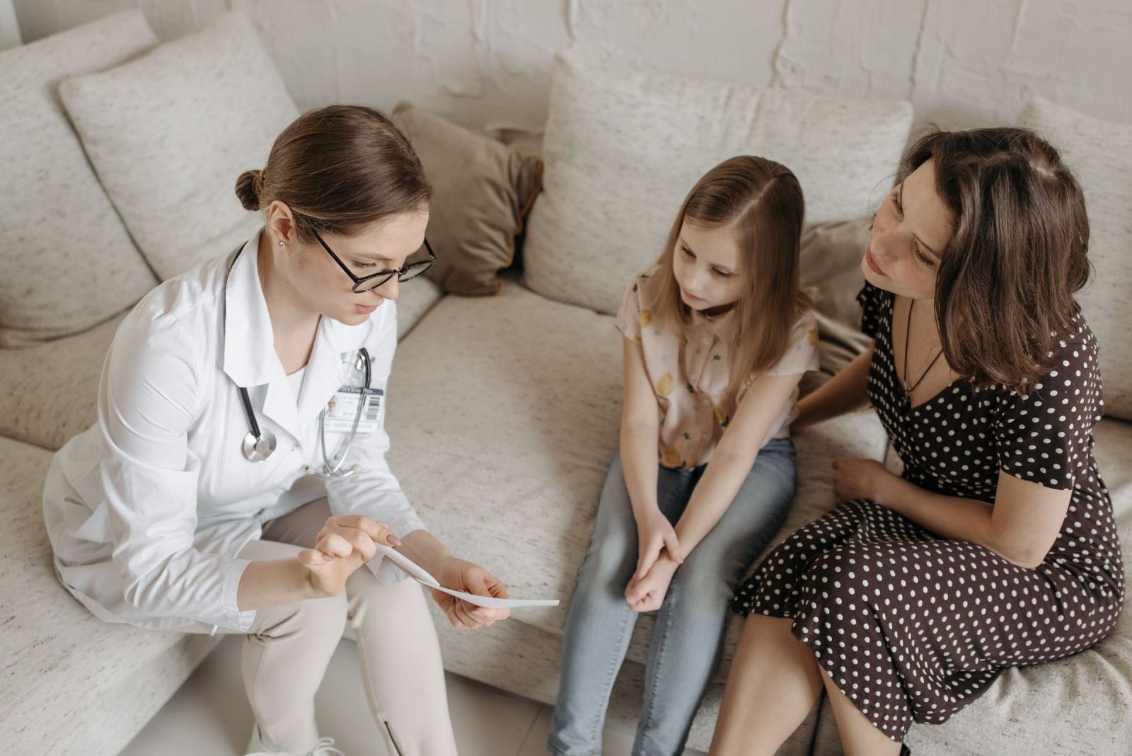 A healthcare professional in a white coat discusses a document with a woman and a child on a couch
