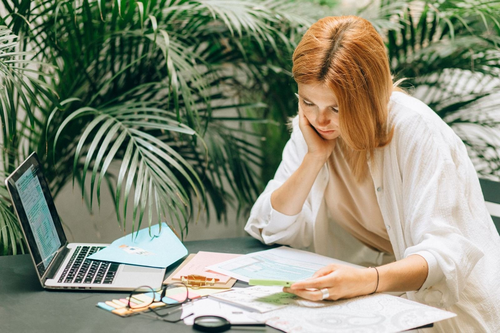 A woman views papers on a desk beside a laptop and houseplants